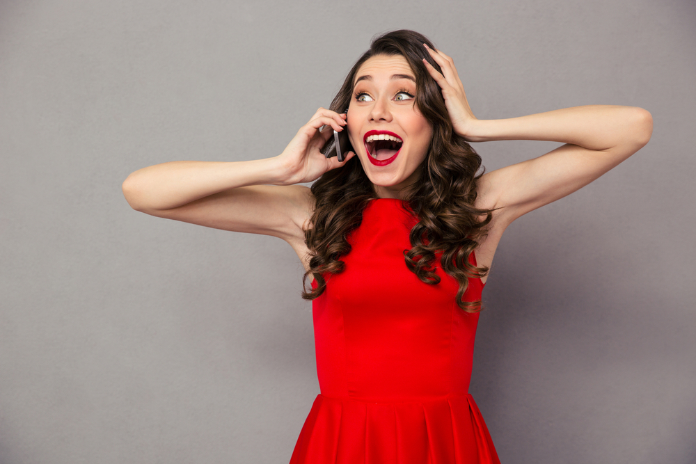 Portrait of a happy amazed woman in red dress talking on the phone over gray background